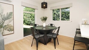 A dining area with a round table, four chairs, a vase with plants, and large windows with a view of greenery. - Early Escape-Welcome to our Eco Friendly and Peaceful Escape in Rural Hamilton! (Christchurch)