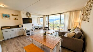 A modern living room with a sofa, a wooden table, and a bench. There is a kitchen area with cabinets and a TV. A large window offers a view of the outdoors. - 27th Fl, 1BR, Beautiful Mountain View by Waikiki Beach, WiFi, AC, Free Parking (Honolulu)