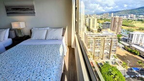 A hotel room with a bed, a nightstand, a lamp, and a view of a cityscape through a large window. - 27th Fl, 1BR, Beautiful Mountain View by Waikiki Beach, WiFi, AC, Free Parking (Honolulu)