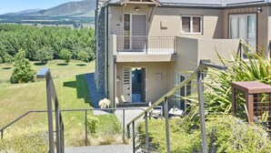 Una casa de dos pisos con balcón, un porche pequeño y vistas a un paisaje verde con árboles y montañas al fondo.