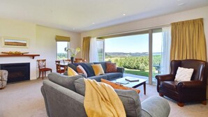 Amplia sala de estar con un sofá gris, un sillón de cuero marrón y una mesa de centro. Cuenta con una chimenea, un comedor con sillas y un gran ventanal que ofrece vistas al exterior.