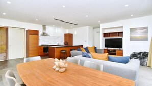 A modern living room with a wooden dining table, grey sofa with colorful cushions, a kitchen area with a stove, and a flat-screen TV mounted on the wall. - High Country Villa 250-Luxury Villa in Terrace Downs (Christchurch)