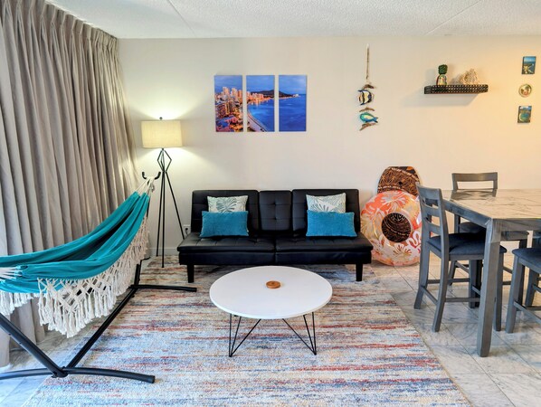 A modern living room with a black sofa, a round white table, a colorful rug, a hammock, and a cityscape painting on the wall. - 22nd Fl, Free parking, AC, WIFI, Beautiful Mountain View (Honolulu)