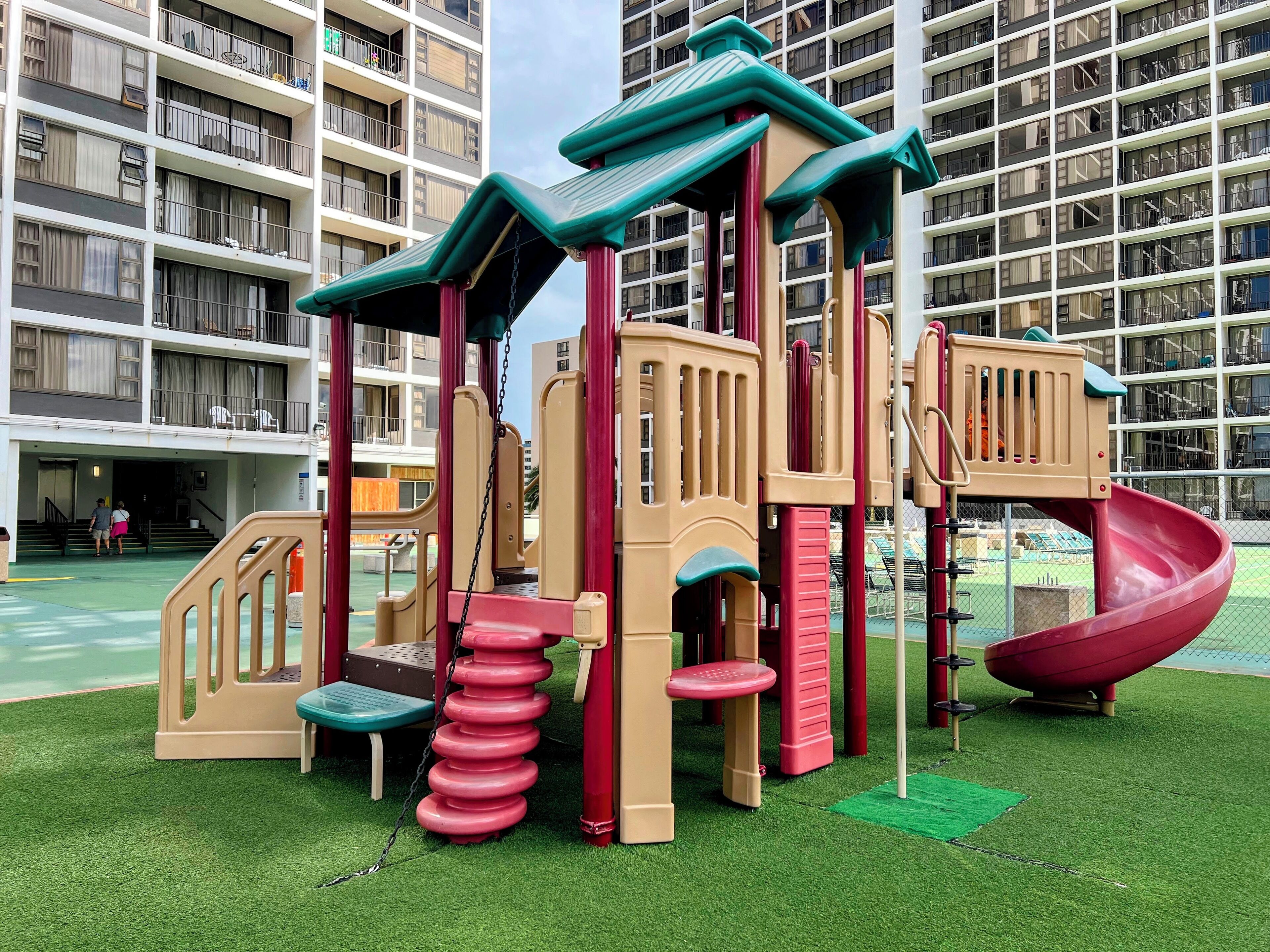 A playground with a slide and climbing structures, surrounded by a green field and a high-rise building in the background.