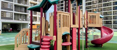 A playground with a slide and climbing structures, surrounded by a green field and a high-rise building in the background.