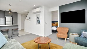 Un salon moderne avec un canapé, un fauteuil et une table basse. On y trouve également un coin cuisine équipé d’un réfrigérateur et d’un luminaire suspendu.