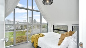 Une chambre avec de grandes fenêtres, un lit recouvert de draps blancs, un plaid jaune et une vue sur la ville.