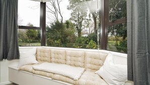 A white cushioned bench with a cushion, positioned in front of a window with a view of greenery and trees. - Narrows Hideaway-Rural retreat near Hamilton Airport (Christchurch)