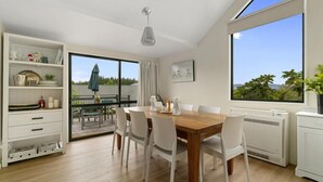 Un coin repas avec une table en bois et des chaises blanches, un meuble blanc avec des étagères, une grande fenêtre donnant sur la verdure et un luminaire au plafond.