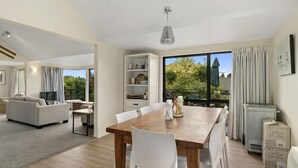 Un salon moderne avec une table à manger en bois, des chaises blanches, un canapé et un téléviseur. Une grande fenêtre, ornée de rideaux, offre une vue sur la verdure à l’extérieur.
