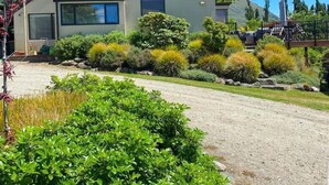 Une maison moderne à deux étages, dotée d’un toit en pente, de grandes fenêtres et d’un jardin bien entretenu où poussent diverses espèces d’arbustes et de plantes.