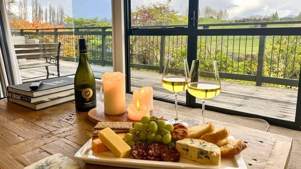 Une table avec un plateau de fromages, des verres à vin, une bougie et une bouteille de vin, disposés sur un balcon offrant une vue sur un paysage verdoyant.