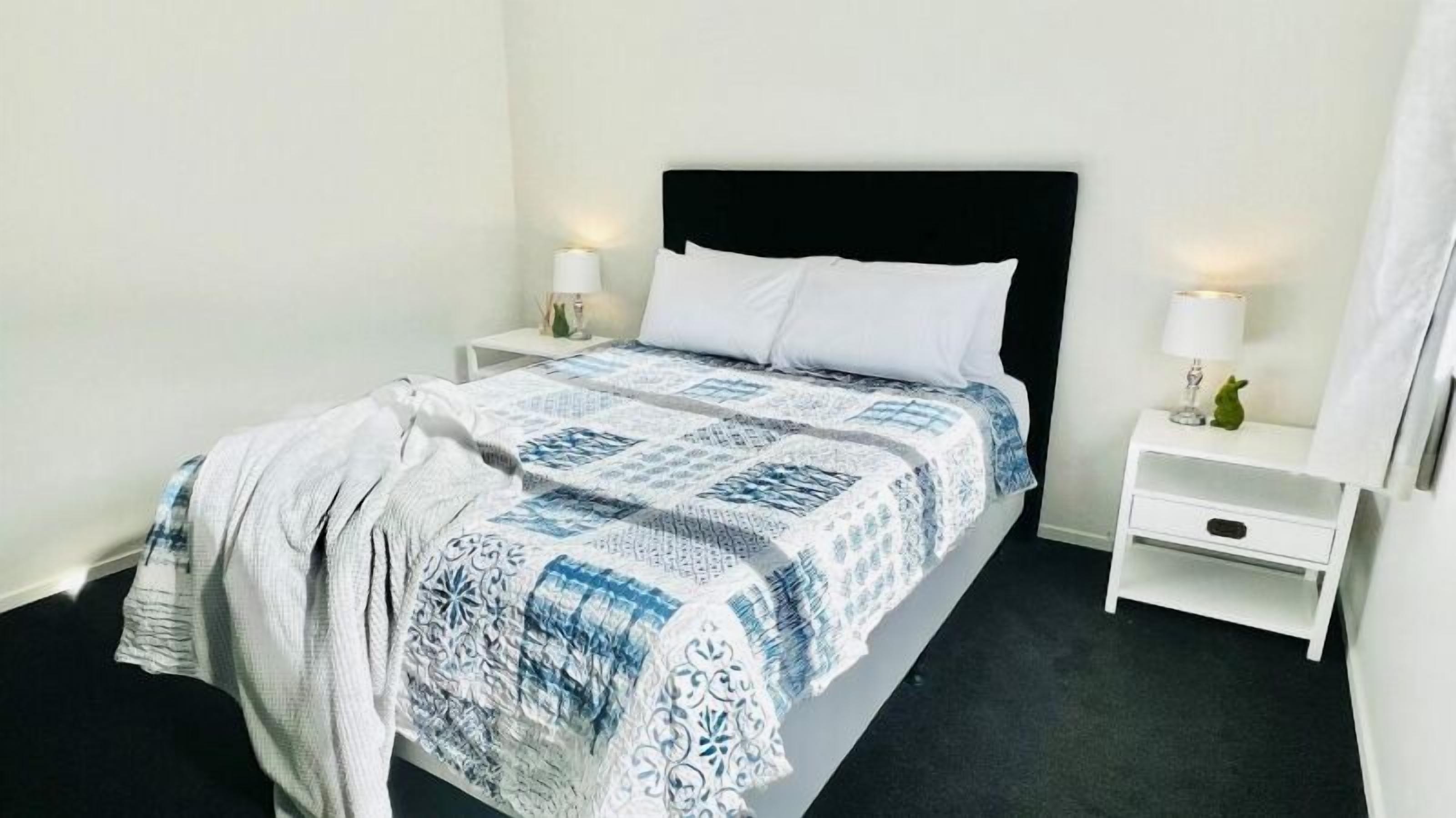 Una cama bien hecha con una colcha estampada y almohadas, dos mesitas de noche con lámparas y una pequeña mesita auxiliar con un objeto decorativo.