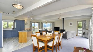 Amplia sala de estar con mesa de madera, sillas, sofá y televisor. Se observa un área de cocina con un armario azul y un ventanal grande.