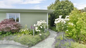 Una casa verde con un sendero que lleva a la puerta principal. Hay plantas con flores blancas y diversas arbustos y árboles alrededor de la casa.