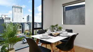 A modern dining area with a table set for four, large windows offering a city view, and a potted plant on the windowsill. - Worcester Terrace 201 - Luxurious City Apartment (Christchurch)