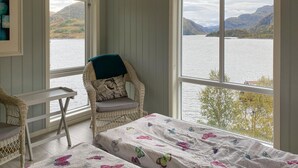 Una habitación con vista a un lago y montañas, una silla de mimbre con cojín, una mesita y una cama con colcha de estampado de mariposas.