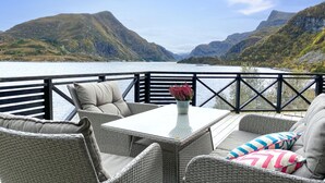 Una terraza con mobiliario de mimbre y una mesa, con vista a un lago y montañas.