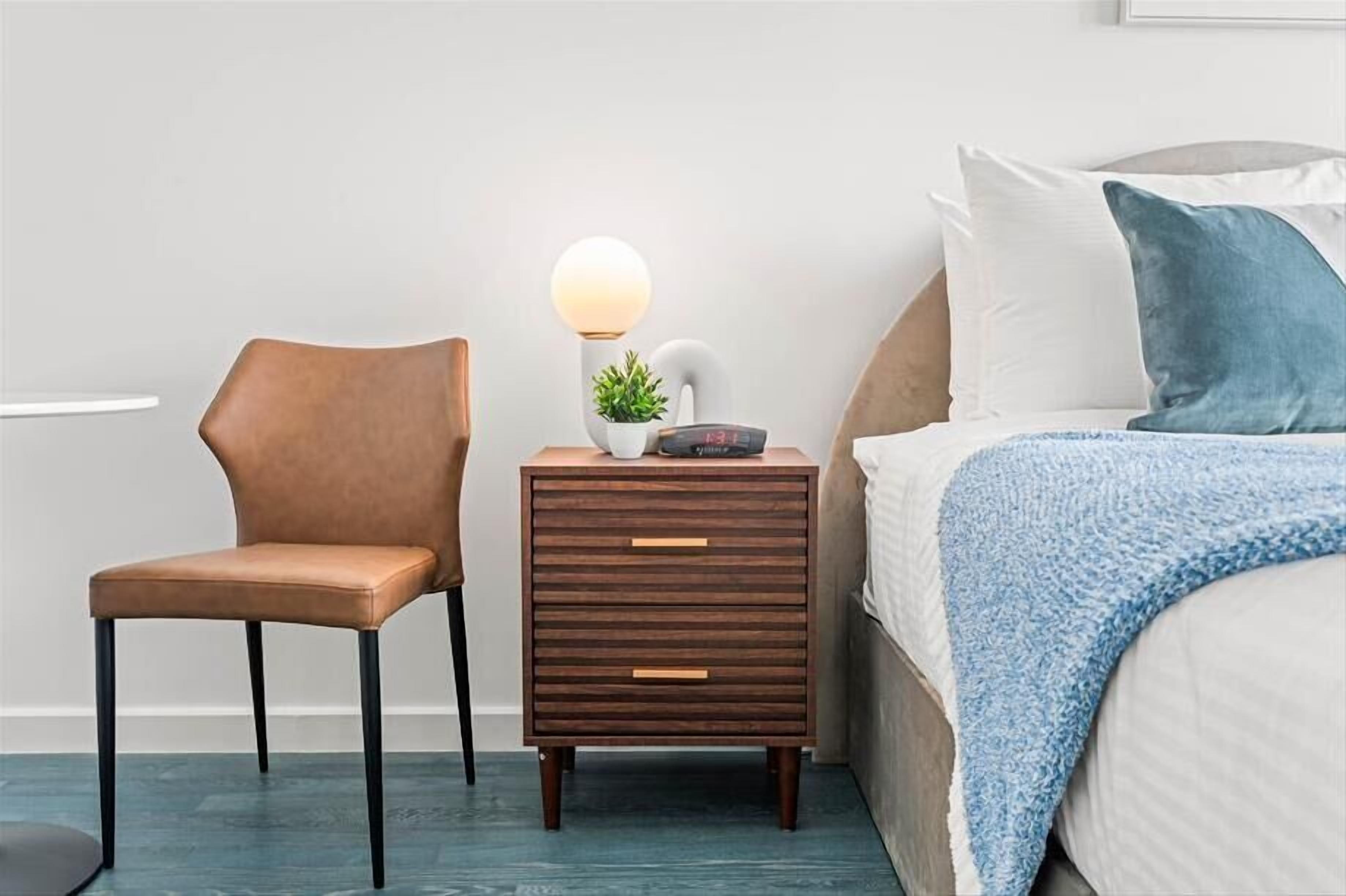 A bedroom with a bed, a bedside table with a lamp, a chair, and a small plant.