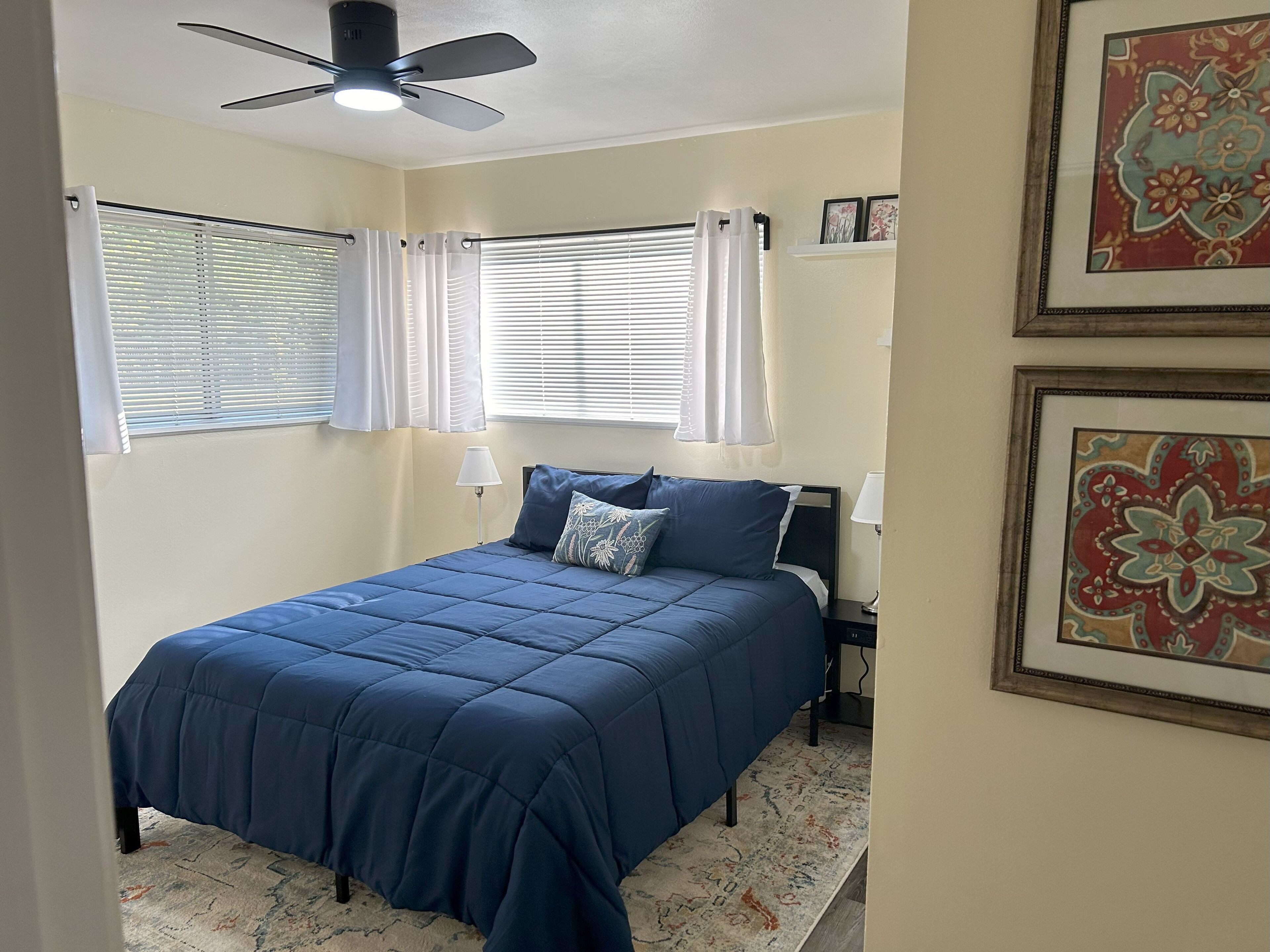 A bedroom with a bed, ceiling fan, window with curtains, bedside lamps, and framed artwork on the wall.