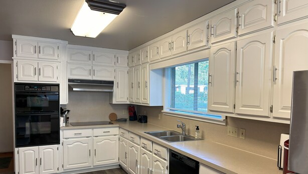 Una cocina moderna con armarios blancos, horno integrado, lavavajillas y ventana con persianas.
