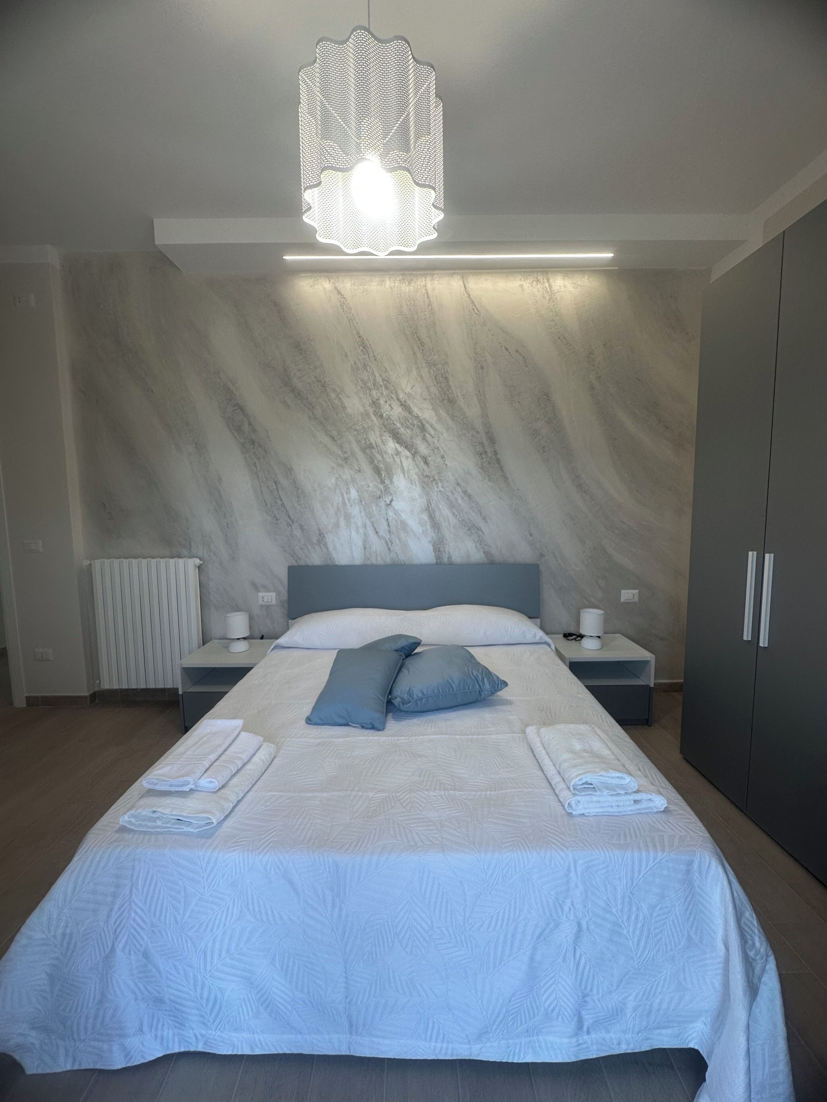 A modern bedroom with a large bed, a marble-patterned wall, a hanging light fixture, and a wardrobe.