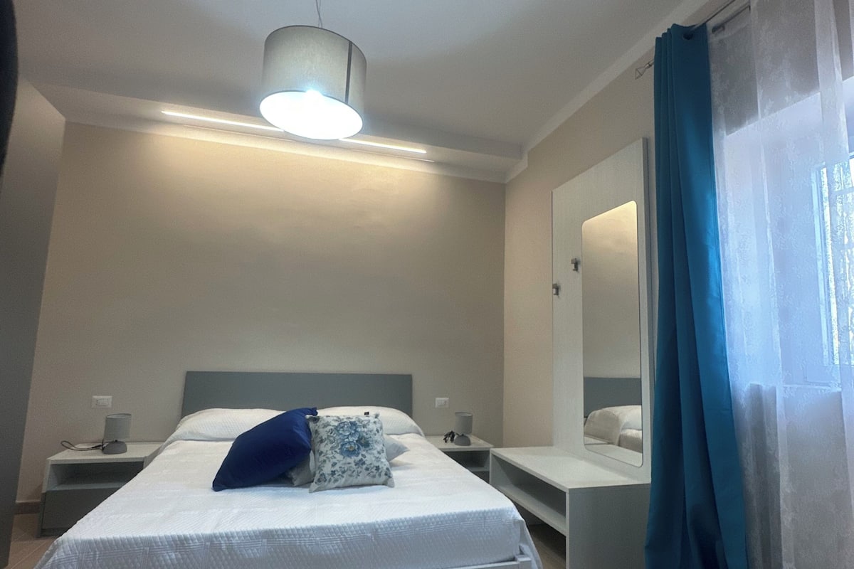 A bedroom with a bed, bedside tables, a mirror, and a hanging light fixture.