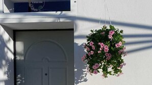 Una puerta blanca con un ventanal circular, una maceta colgante con flores rosas, y un letrero con texto sobre la puerta.
