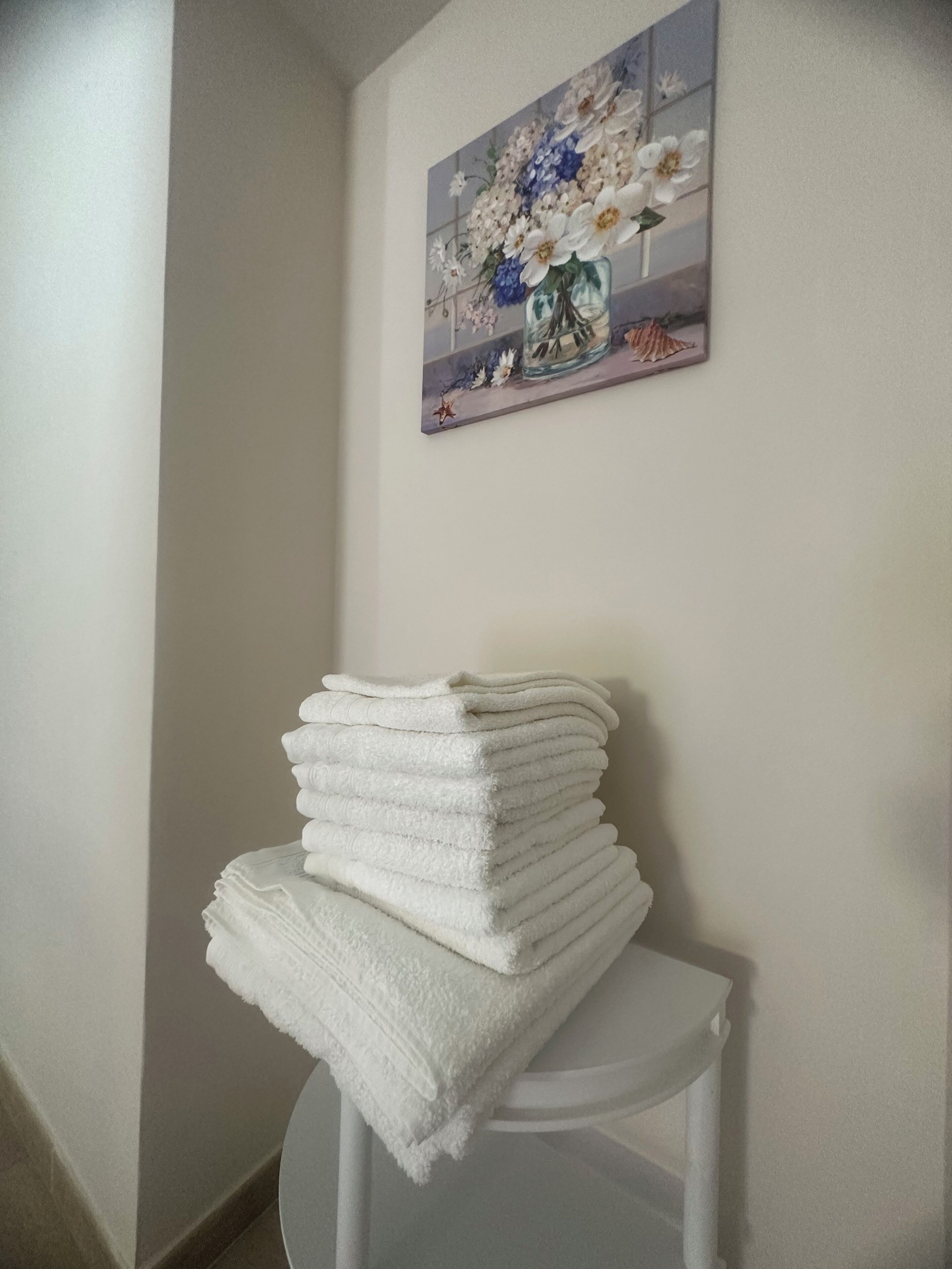 A stack of white towels on a white chair against a plain wall with a floral painting above it.