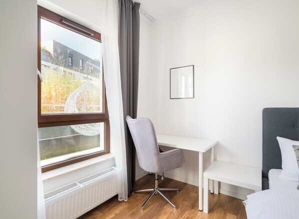 Une chambre d’hôtel moderne avec un bureau blanc, une chaise grise, un lit avec une literie blanche, un tableau encadré au mur et une fenêtre donnant sur des immeubles et des arbres.