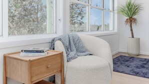 Una habitación con un ventanal grande, una mesita de noche con libros, una silla y una planta en maceta.