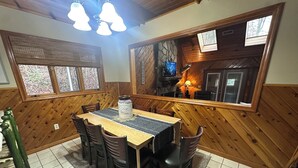 Zona pranzo con tavolo e sedie in legno, un tappeto e un vaso decorativo. Sono presenti finestre con vista sull'esterno e un lampadario.