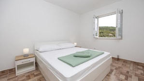 Un dormitorio minimalista con una cama individual, una mesita de noche, una lámpara de noche y una ventana con vistas a la vegetación.