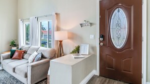 A living room with a beige sofa, a lamp, a framed certificate, and a brown door with a glass panel. - Serenity Villa Hermosa, Jacuzzi, Arcade & Fun Games, Backyard, Outdoor Furniture (Moreno Valley)