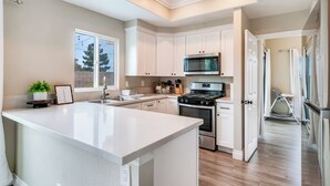 A modern kitchen with white cabinetry, a central island, stainless steel appliances, and a view of the outdoors through a large window. - Serenity Villa Hermosa, Jacuzzi, Arcade & Fun Games, Backyard, Outdoor Furniture (Moreno Valley)
