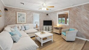 A spacious living room with a white sofa, a white coffee table, a wicker armchair, a ceiling fan, and a flat-screen TV. - Tiki Tides OBX 4-Bedroom Beach Box in Kill Devil Hills, Steps from the Beach! (Kill Devil Hills)