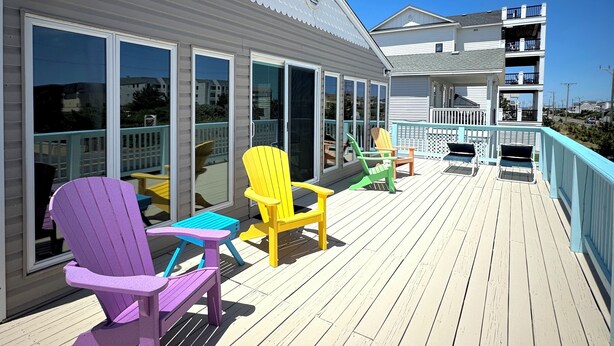 A wooden deck with colorful Adirondack chairs and a building with large windows. - Tiki Tides OBX 4-Bedroom Beach Box in Kill Devil Hills, Steps from the Beach! (Kill Devil Hills)