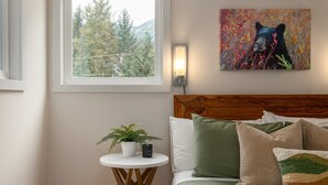 A bedroom with a wooden headboard, a bed with multiple pillows, a small round table with a plant, a wall-mounted lamp, and a window with a view of trees and mountains. - 1BR | Gondola Village | Near Gondola | Free Parking | Updated Unit | Incredible Location (Whistler)