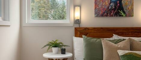 A bedroom with a wooden headboard, a bed with multiple pillows, a small round table with a plant, a wall-mounted lamp, and a window with a view of trees and mountains.