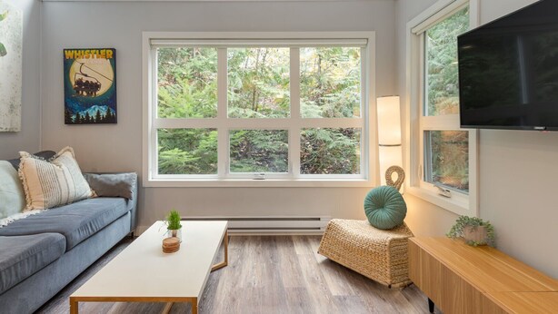 A cozy living room with a large window, a flat-screen TV, a sofa, a coffee table, and a small side table with a plant. - 1BR | Gondola Village | Near Gondola | Free Parking | Updated Unit | Incredible Location (Whistler)