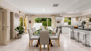 A spacious dining area with a glass table and white chairs, a kitchen with modern appliances, and a view of greenery outside. - StayAU Expansive 7BR Villa Warrandyte (Warrandyte)