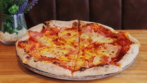 Una pizza sobre un plato redondo, una mesa de madera y un florero con flores al fondo.