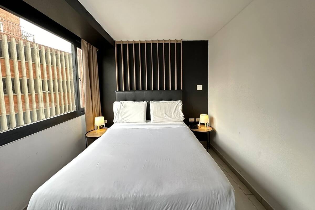 Une chambre d’hôtel moderne avec un grand lit, deux tables de chevet avec des lampes, une tête de lit sombre et une fenêtre avec des stores.
