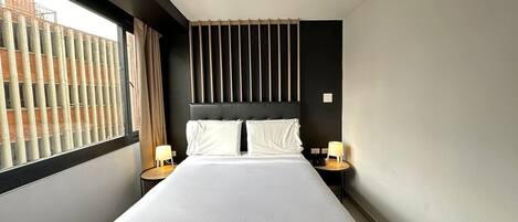 Ein modernes Hotelzimmer mit einem großen Bett, zwei Nachttischen mit Lampen, einem dunklen Kopfteil und einem Fenster mit Jalousien.