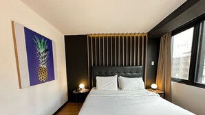 Una habitación de hotel con una cama grande, cabecera con listones verticales, mesita de noche con lámpara, un cuadro con motivo de piña en la pared y una ventana con cortinas.