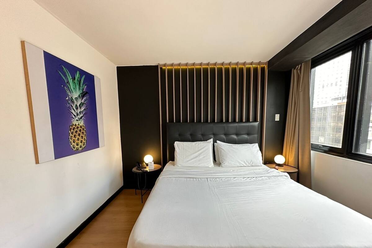 Une chambre d’hôtel avec un grand lit, une tête de lit à lattes verticales, une petite table de chevet avec une lampe, une œuvre d’art représentant un ananas encadrée au mur et une fenêtre avec des rideaux.