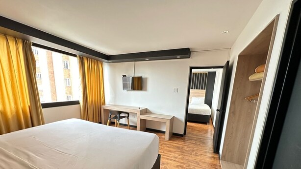 A hotel room with a large bed, a desk, and a chair. There is a TV mounted on the wall and a door leading to another room. - Hotel San Francisco de Asis (Bogotá)