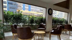 Un área de estar interior moderna con sillones de cuero y una mesa de madera, ventanas amplias con vistas a un edificio y vegetación.