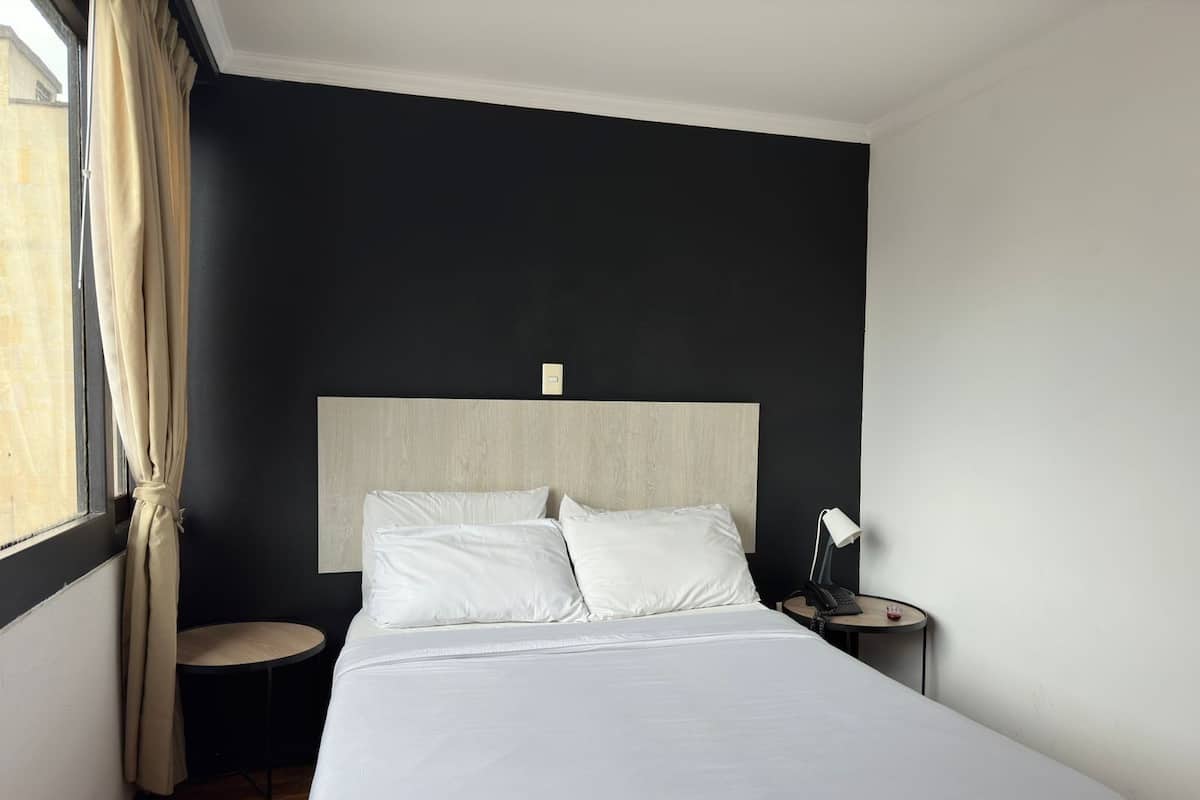 Une chambre d’hôtel avec un grand lit, une table de chevet avec une lampe, une petite table ronde et une fenêtre avec des rideaux.
