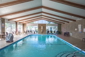 Une piscine intérieure offrant une vue dégagée sur l’extérieur grâce à de grandes fenêtres, des poutres en bois au plafond et plusieurs espaces de détente aménagés autour de la piscine.